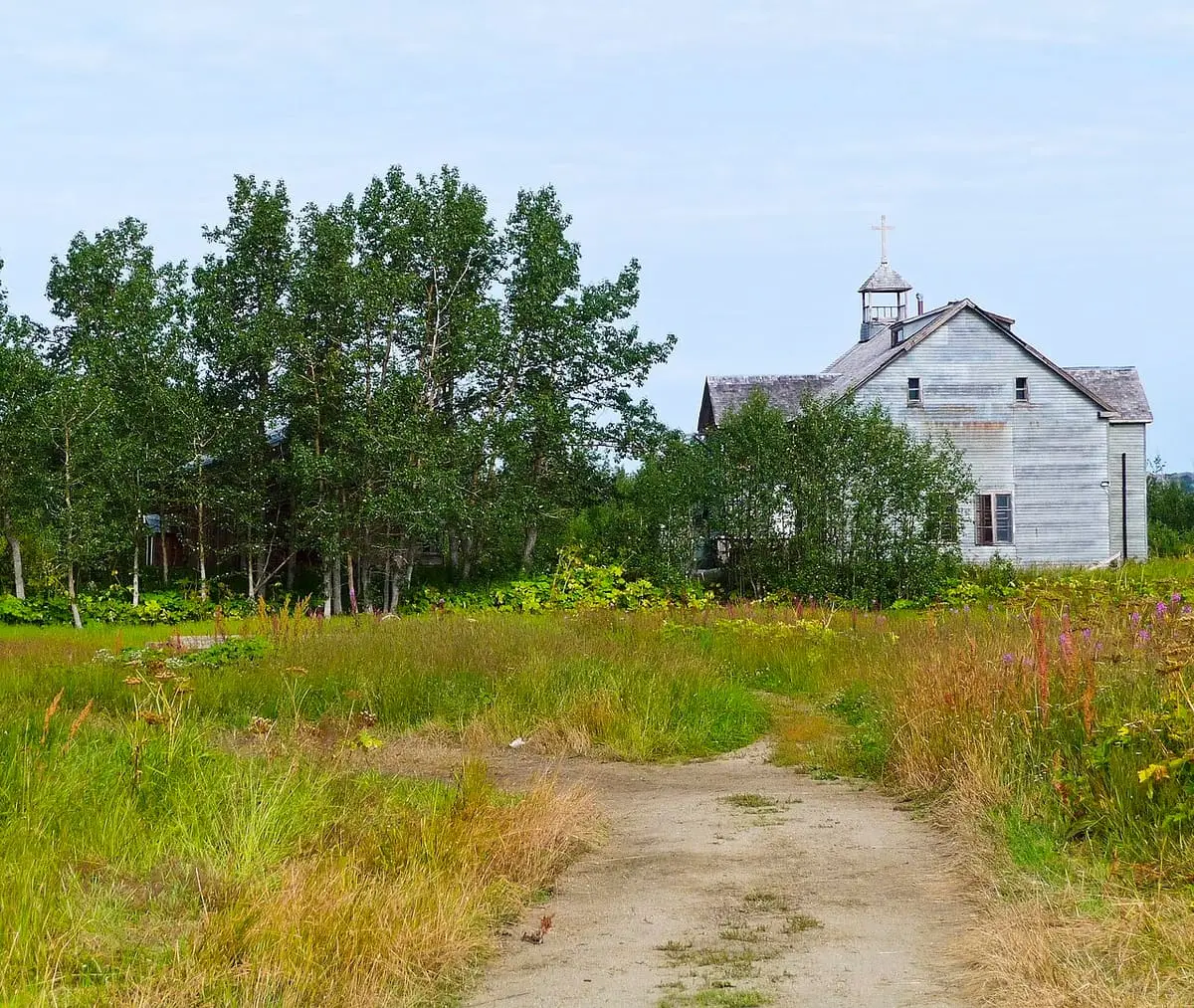 Weathered white church at Pilgrim Hot Springs beyond a grassy track and stand of trees