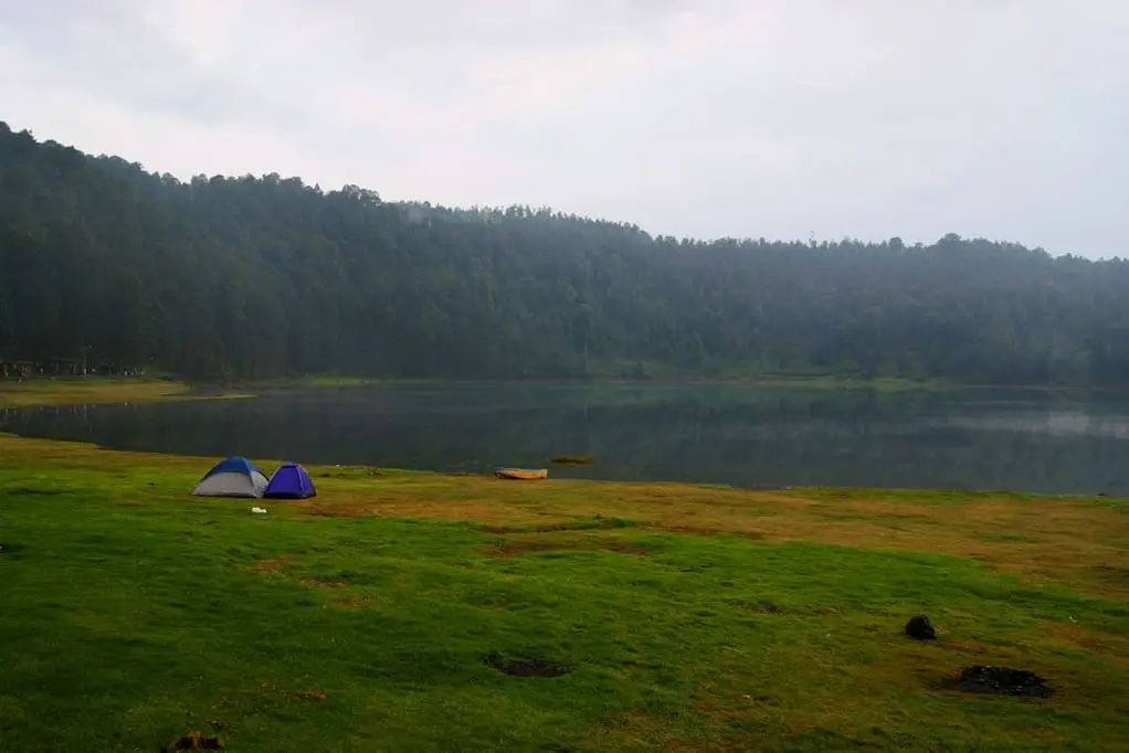 Tents on the grassy lakeshore at Laguna Larga beneath forested hills in Los Azufres