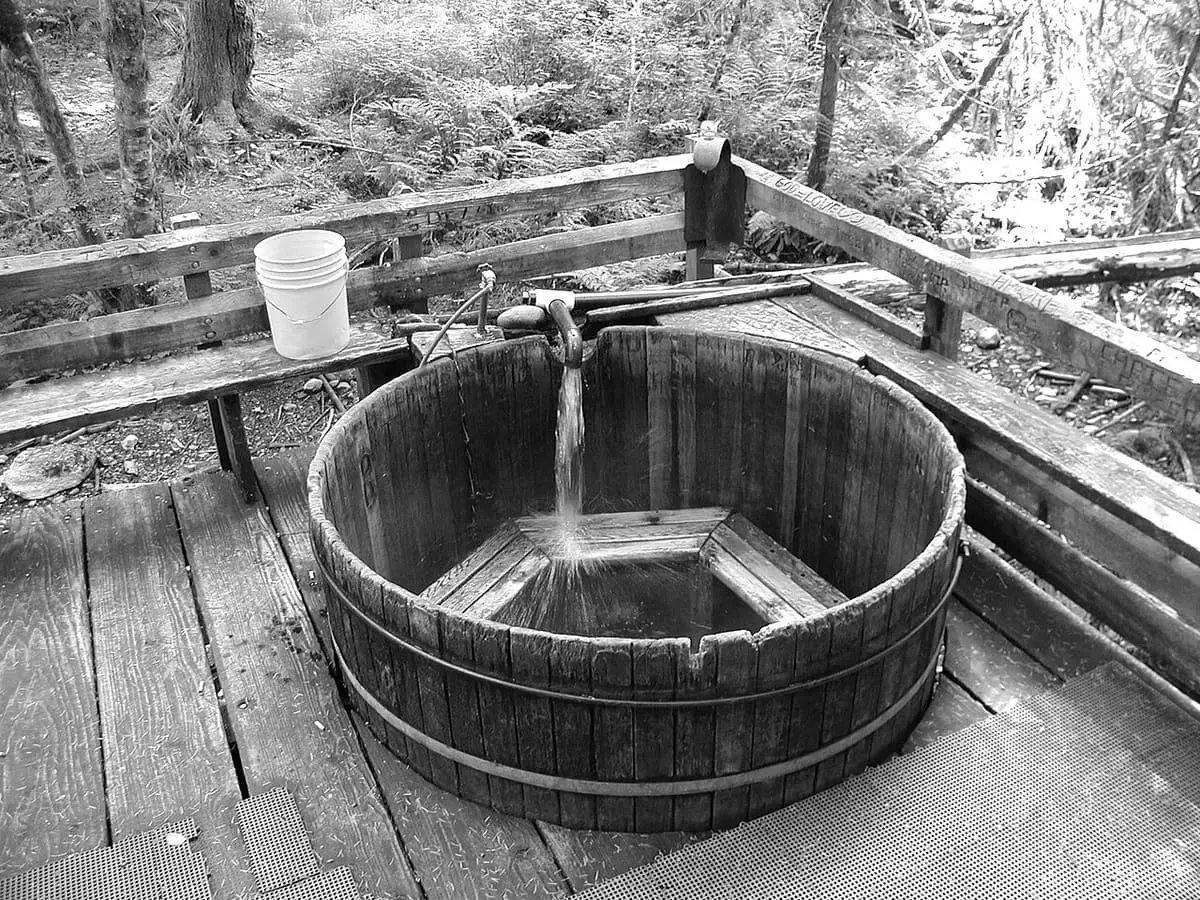 Black and white view of a round wooden soaking tub fed by a log flume at Bagby Hot Springs