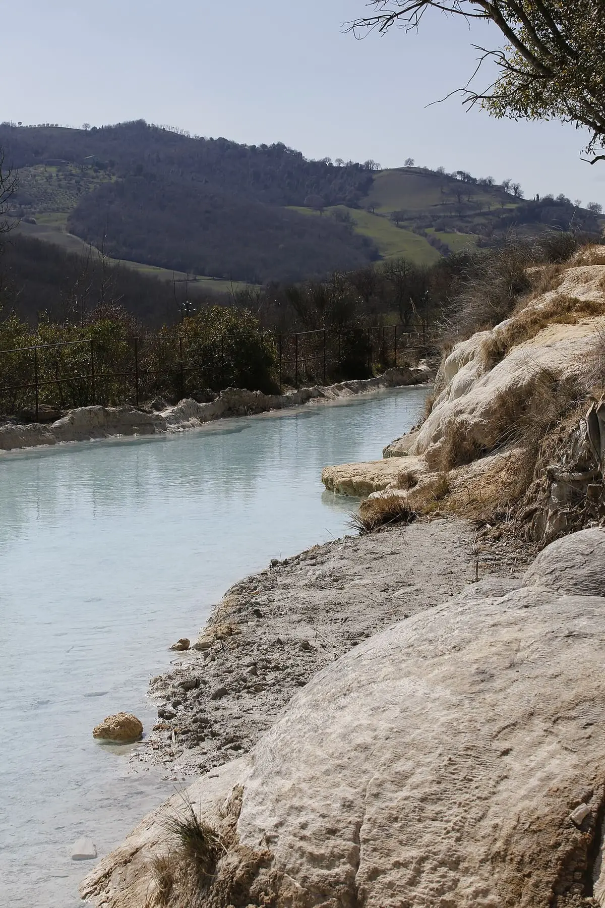 Milky blue thermal pool below white travertine banks at the foot of Bagno Vignoni