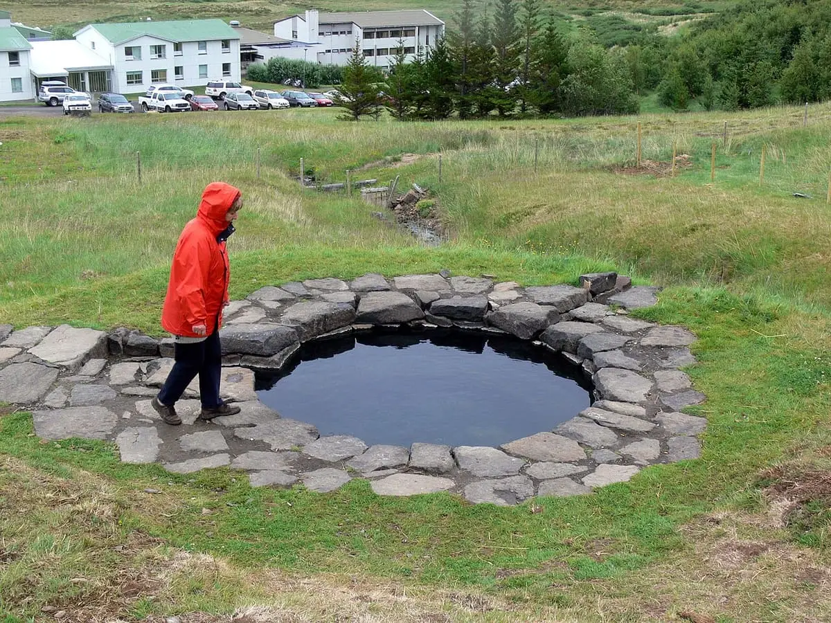 Circular stone rimmed Guðrúnarlaug hot pool in the grass with the hotel buildings at Laugar behind