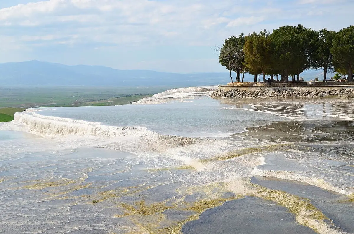 Shallow travertine pools at Pamukkale with trees on the rim and mountains beyond
