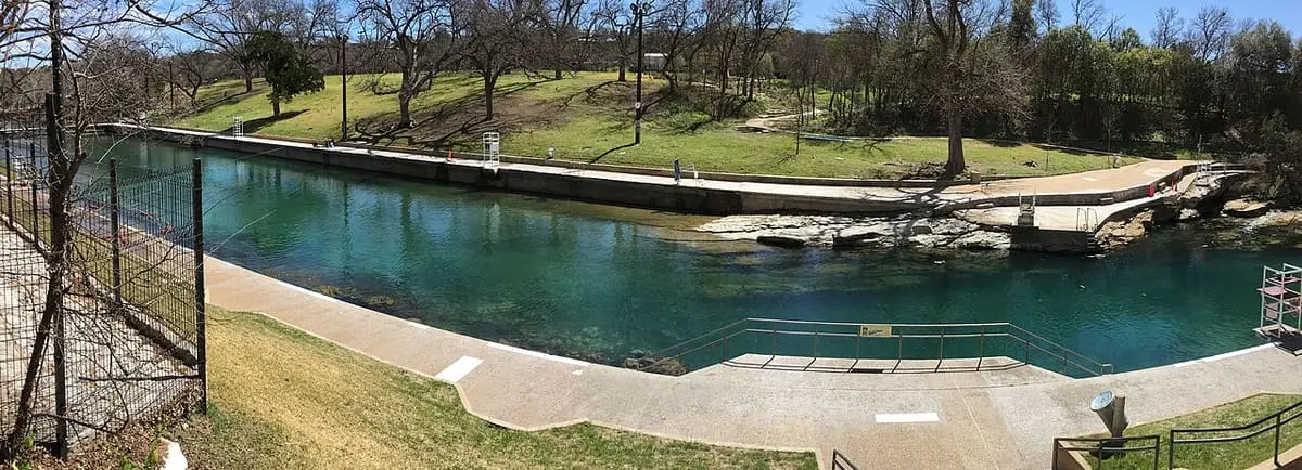 Panoramic view of turquoise Barton Springs Pool beside grassy lawns and leafless trees in Zilker Park