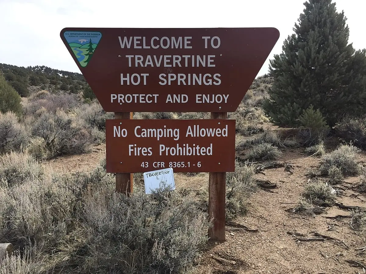 Brown welcome sign for Travertine Hot Springs among sagebrush and pine trees