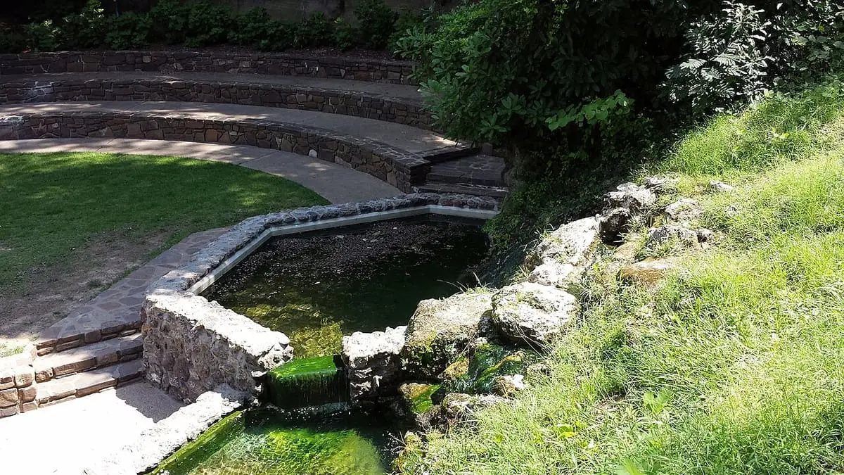 Stone edged pool of green thermal water below curved stone terraces and a lawn