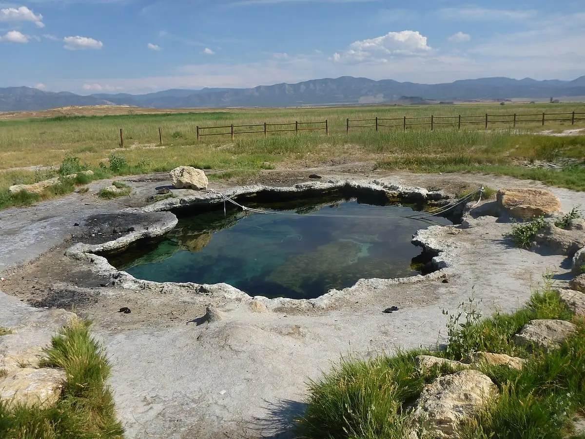 Small round travertine pool at Meadow Hot Springs in grassland with mountains on the horizon