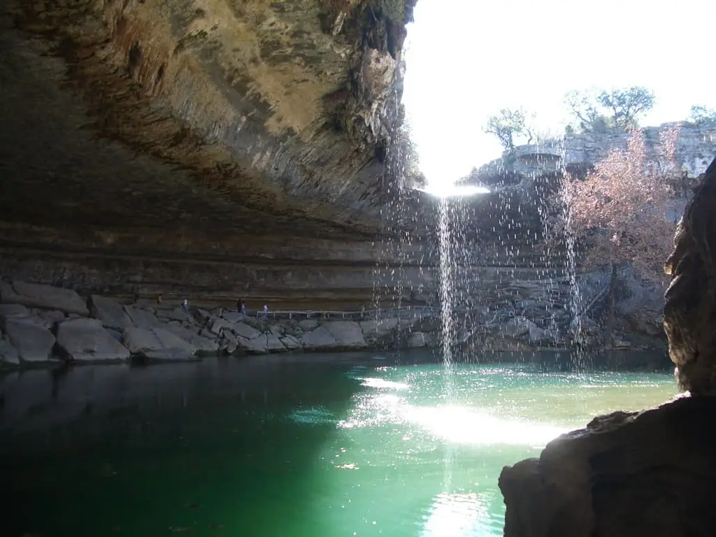 Thin waterfall falling into the sunlit green pool inside the Hamilton Pool grotto