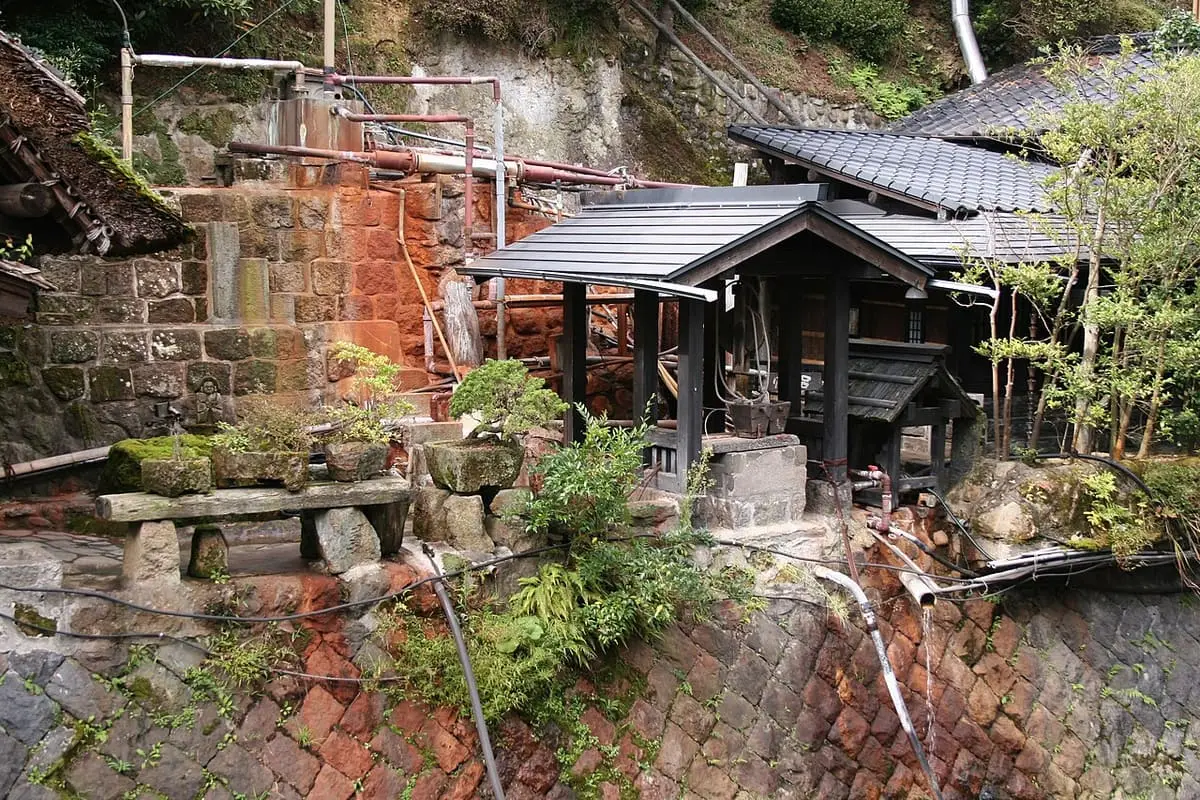 Small wooden bath hut with hot spring pipes on a rust stained stone bank at Kurokawa Onsen