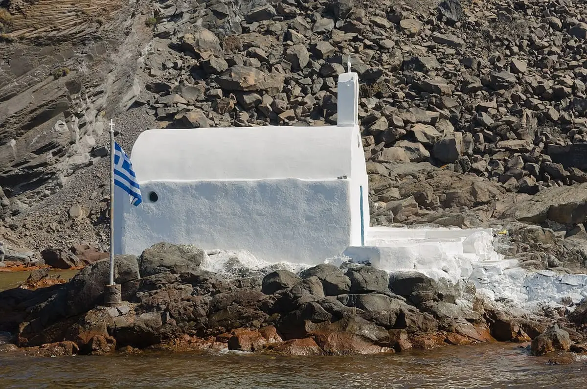 Small whitewashed chapel with Greek flag on the rocky volcanic shore of Palea Kameni