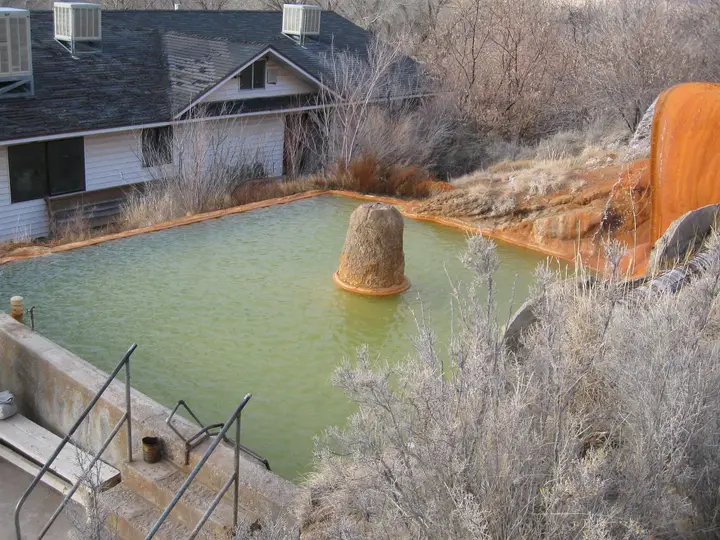 Green mineral pool at Mystic Hot Springs beside orange travertine mounds and frosted brush