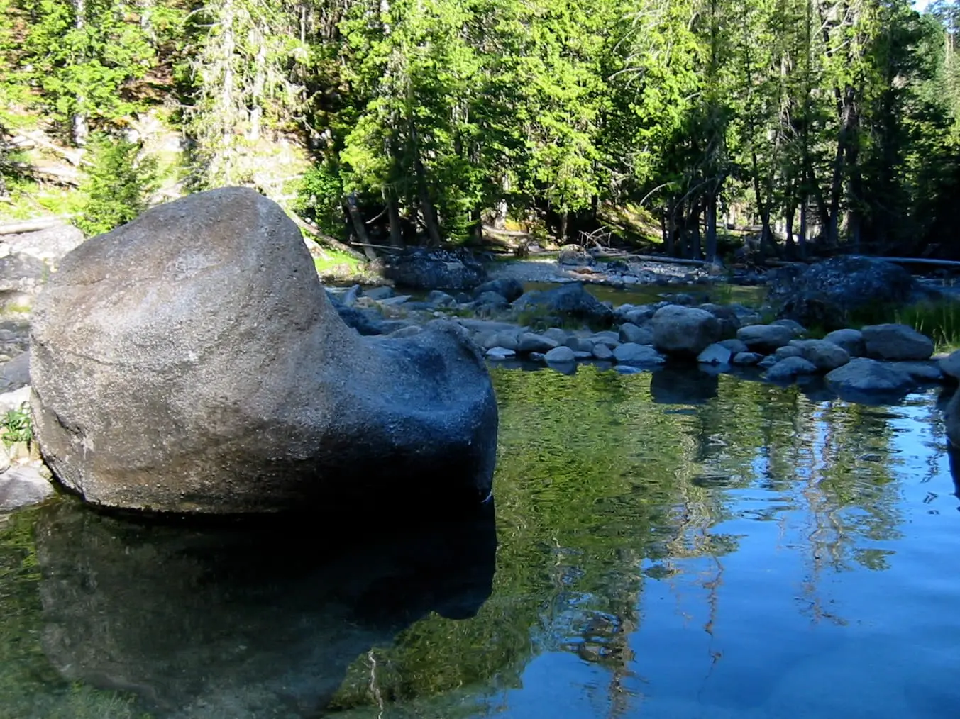 Clear pool at Jerry Johnson Hot Springs with a large boulder, smooth stones and pine forest behind