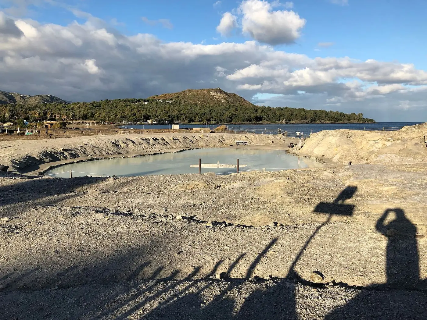 Pale grey mud pool of the Laghetti di Fanghi on Vulcano with wooded hills and the bay behind