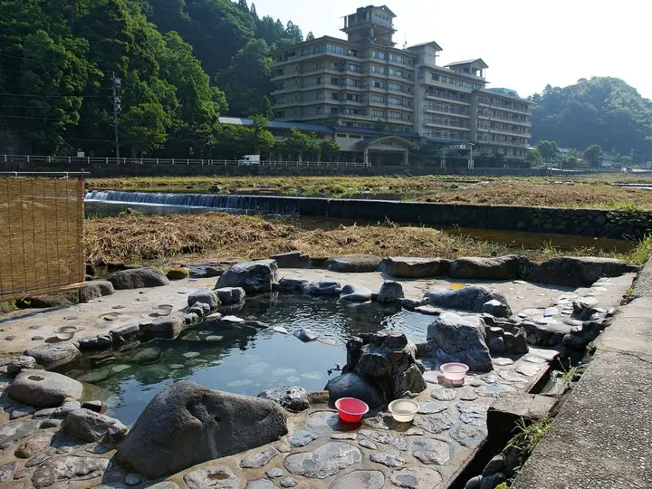 Rock lined Kawara buro open air bath beside the Mitoku River with a large hotel behind at Misasa Onsen