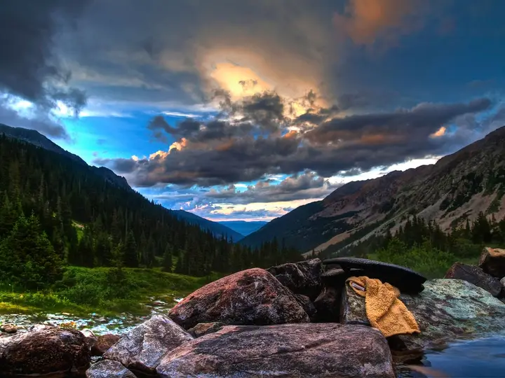 Granite boulders at the edge of Conundrum Hot Springs below a forested valley under dramatic clouds