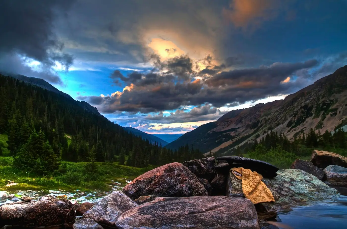 Granite boulders at the edge of Conundrum Hot Springs below a forested valley under dramatic clouds