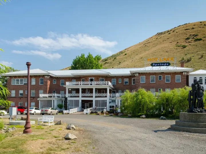 Brick main building of Hot Lake Springs Resort below a dry grassy hillside with a bronze statue out front