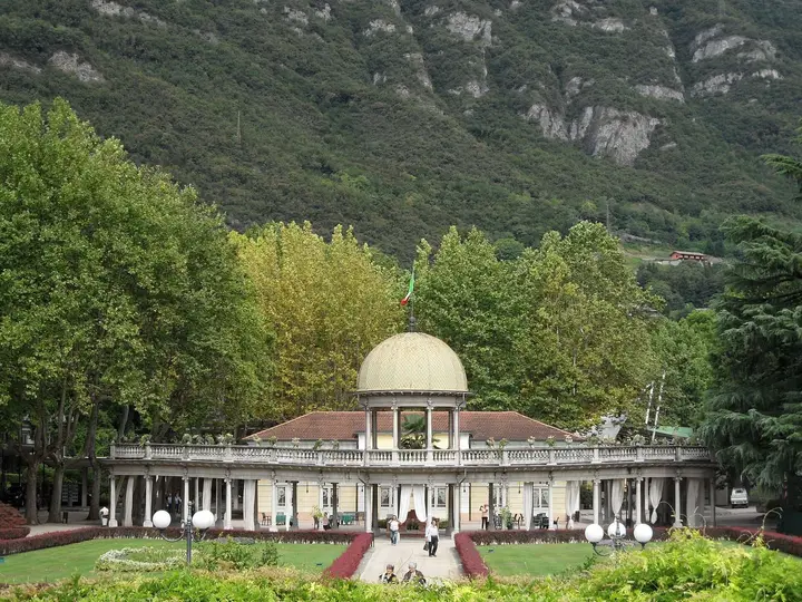 Domed spa pavilion of Terme di Boario in a leafy park below rocky mountains