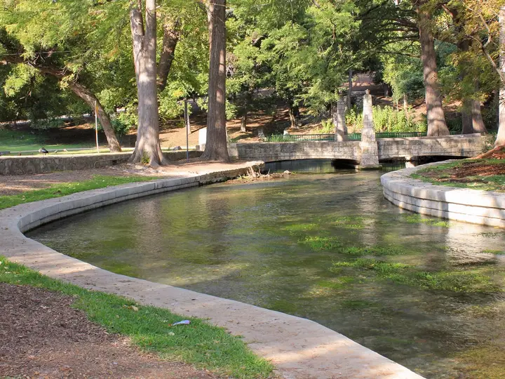 Clear spring fed Comal River channel and stone footbridge under shade trees in Landa Park