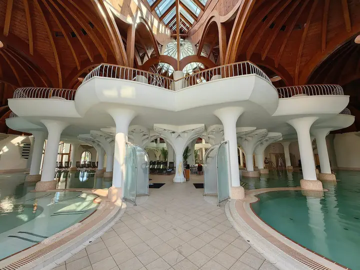 Curving thermal pools beneath white organic columns and a timber dome inside Hagymatikum in Makó