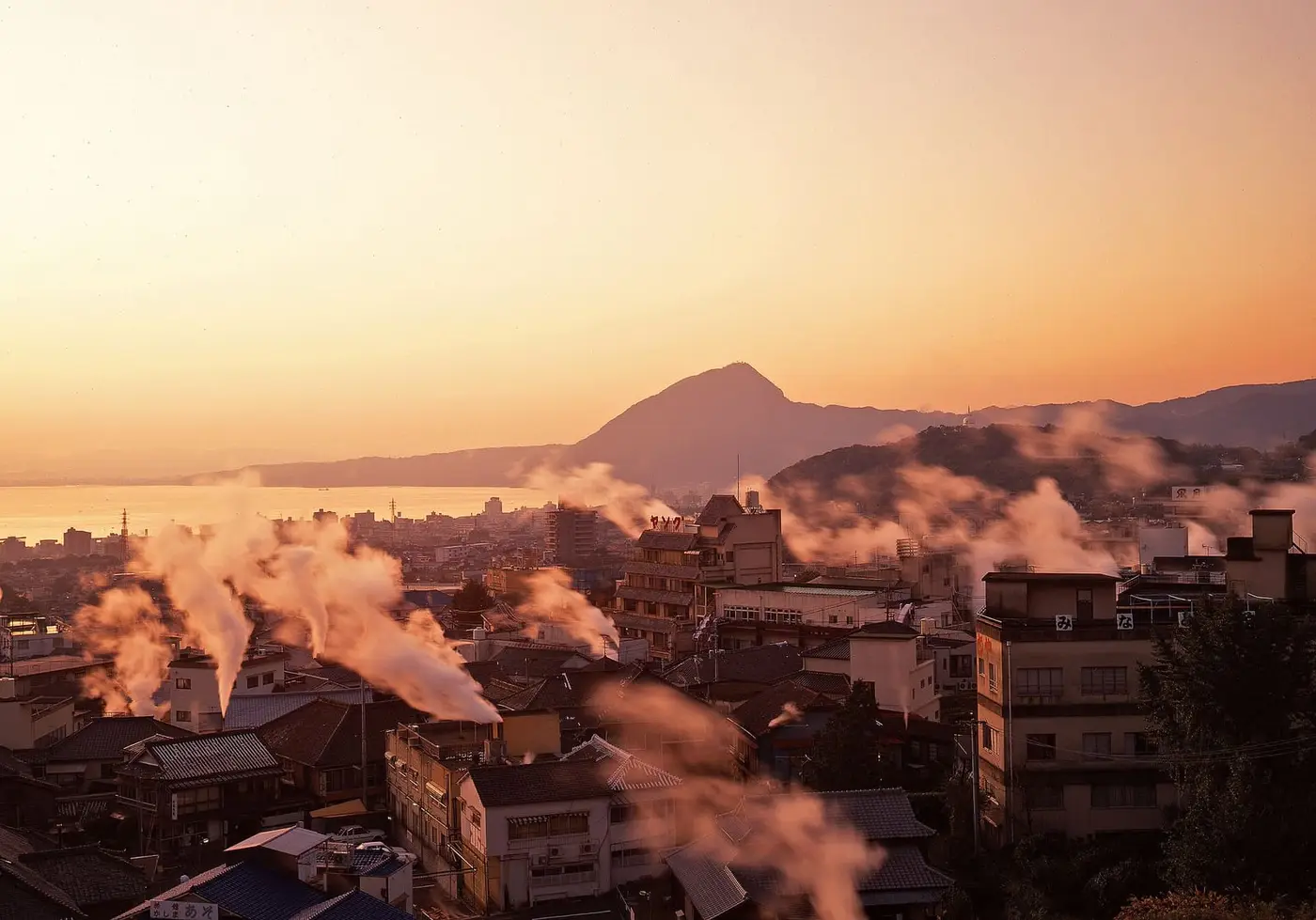 Plumes of hot spring steam rising over Beppu rooftops at sunset with mountains behind