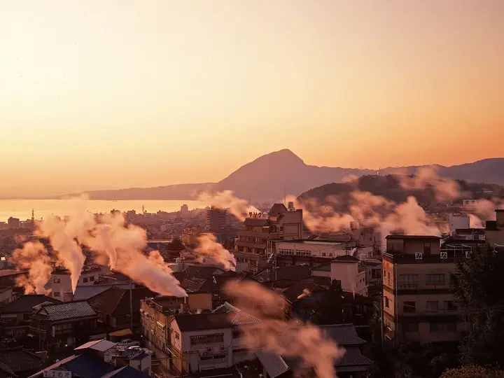 Plumes of hot spring steam rising over Beppu rooftops at sunset with mountains behind