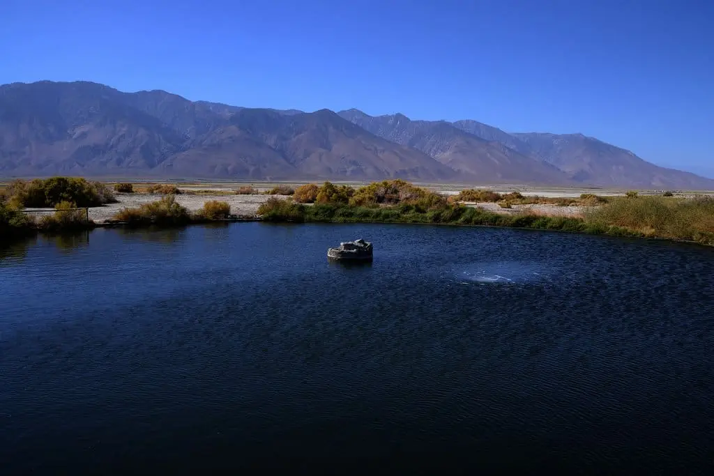Dark blue pool of Dirty Socks Hot Spring with a concrete wellhead, desert brush and bare mountains beyond