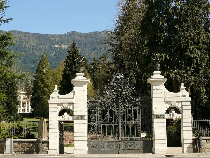 Ornate white gateway to the spa park of Terme di Levico, with forested hills behind