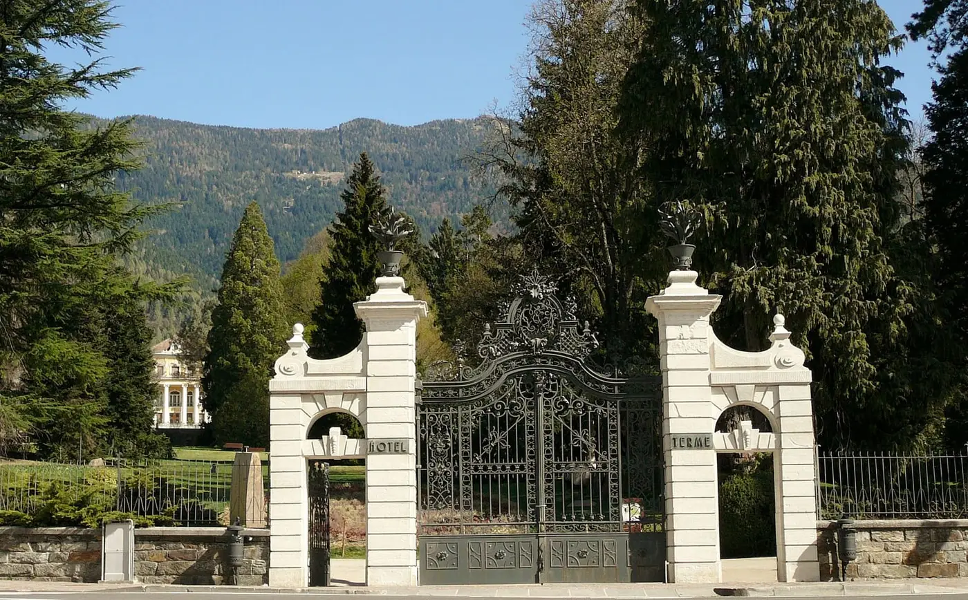 Ornate white gateway to the spa park of Terme di Levico, with forested hills behind