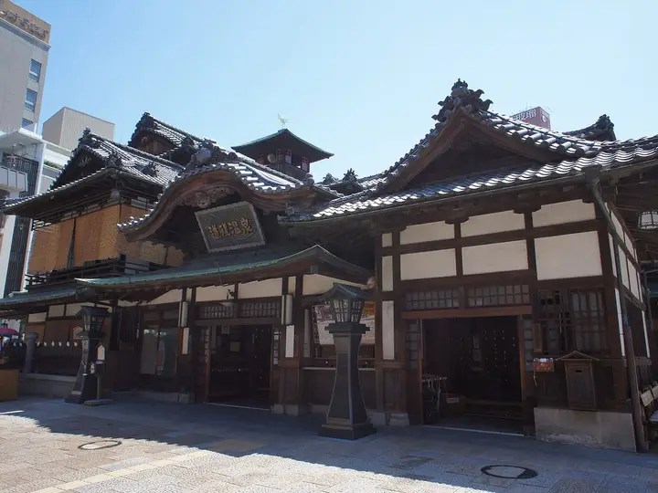 Wooden multi roofed facade of Dogo Onsen Honkan bathhouse under a clear blue sky