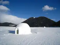 Snow igloo on frozen Lake Shikaribetsu below a dark forested mountain and blue sky