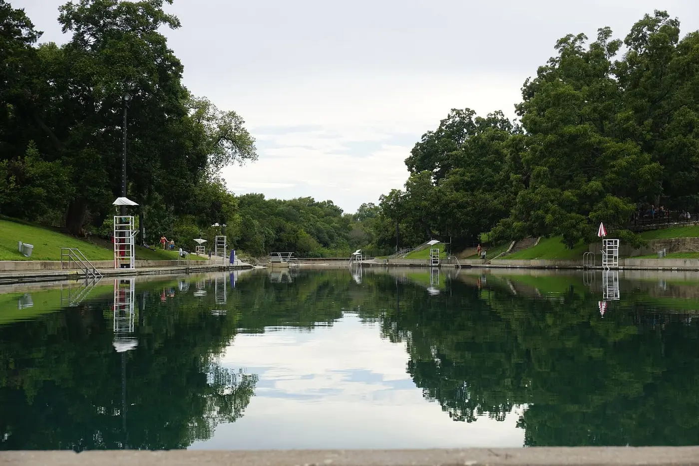 Still green water of Barton Springs Pool reflecting trees and lifeguard stands on a calm day