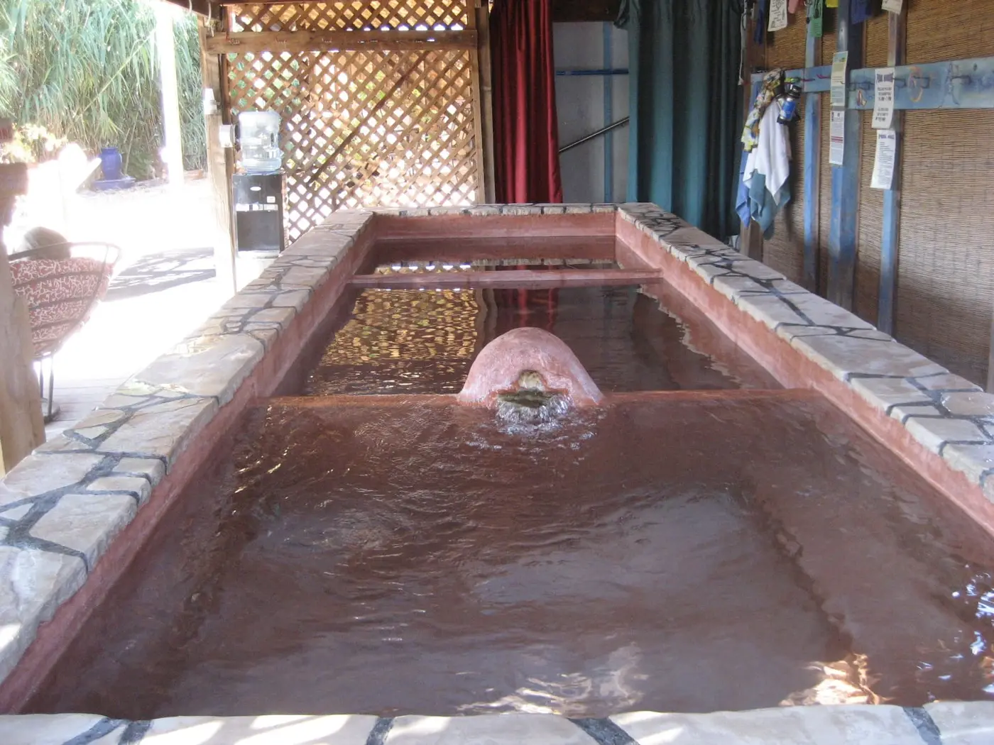Long stone rimmed soaking pool filled with reddish mineral water under a covered pavilion at Riverbend Hot Springs
