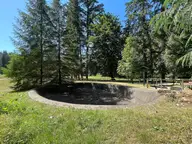 Empty concrete basin at the Flaming Geyser site on a sunny lawn ringed by tall evergreens