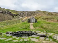 Round stone Guðrúnarlaug pool with a small stone hut on the green hillside behind