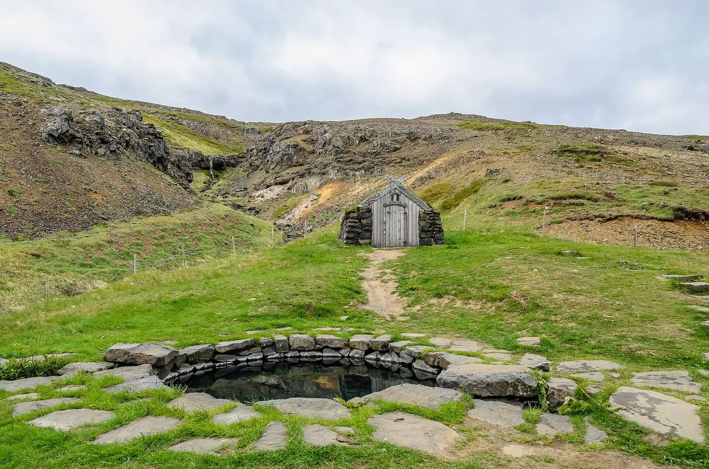 Round stone Guðrúnarlaug pool with a small stone hut on the green hillside behind