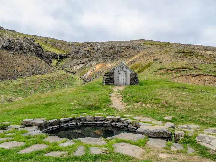 Round stone Guðrúnarlaug pool with a small stone hut on the green hillside behind