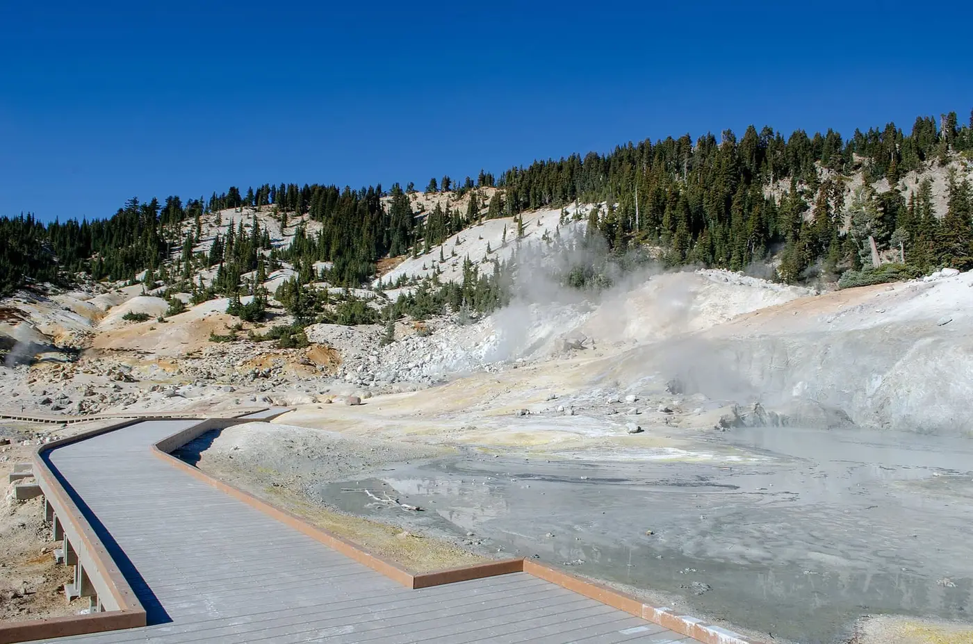 Bumpass Hell boardwalk passing a gray boiling pool with steam rising under a deep blue sky